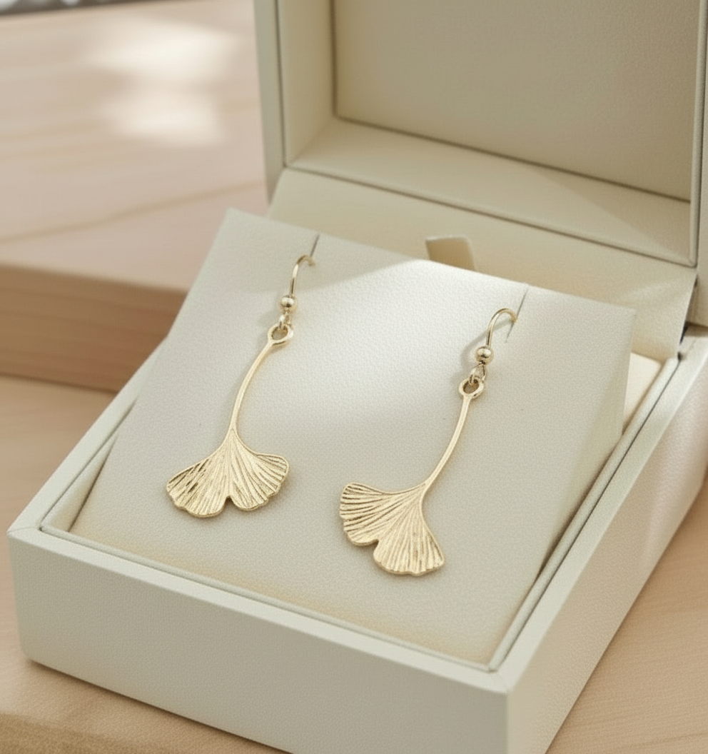 Gold ginkgo leaf dangle earrings in a jewelry box on a light wooden surface