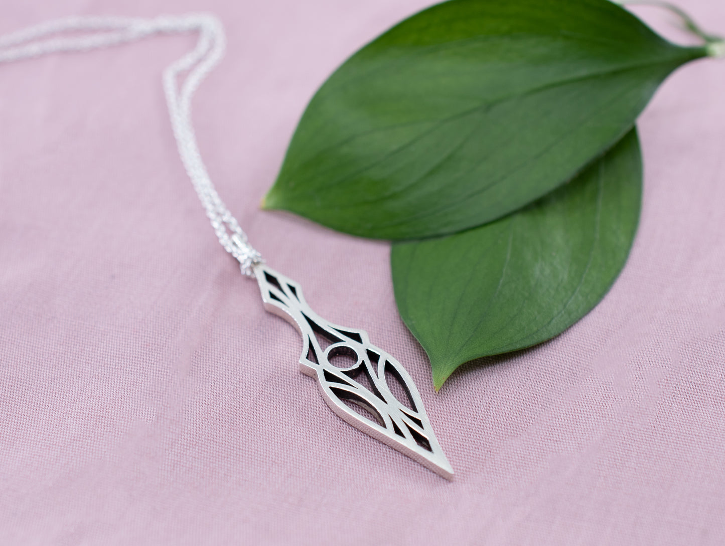 Sterling silver geometric art deco dagger pendant necklace on a pink background with leaves