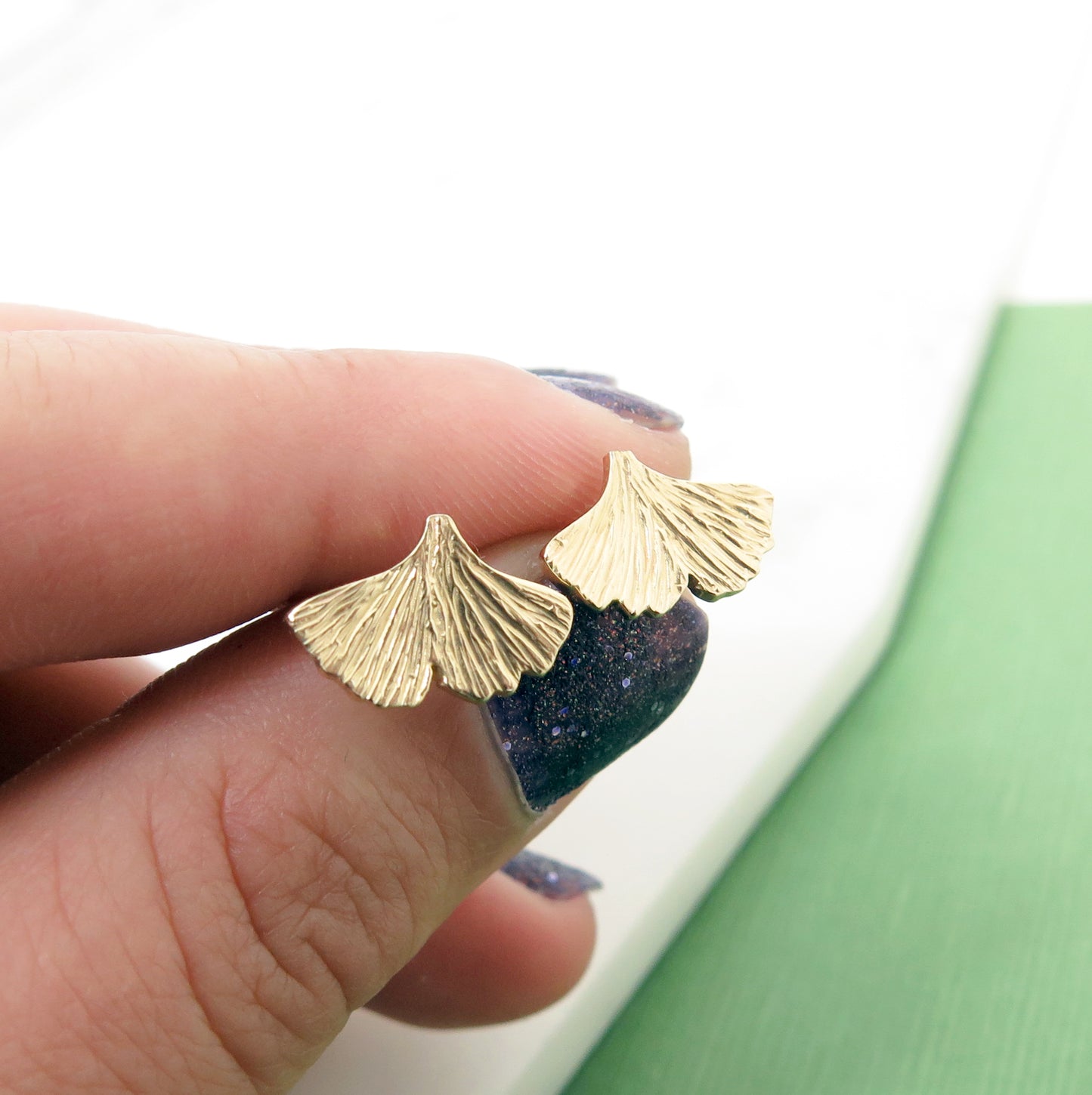 Textured gold ginkgo leaf stud earrings in a hand with nail polish on the nails.