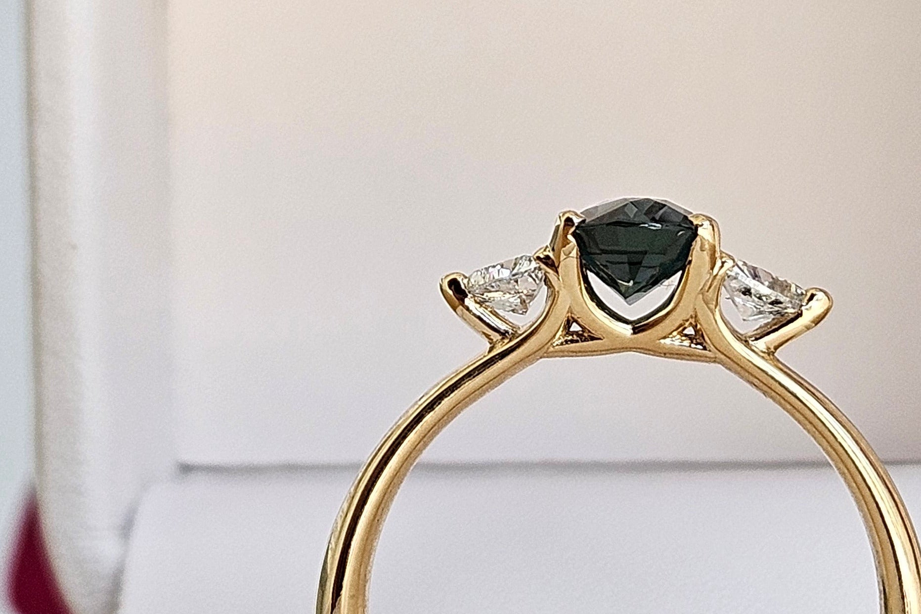 A gold ring with a dark green center sapphire and small clear side stones, displayed inside an open cream-colored ring box.