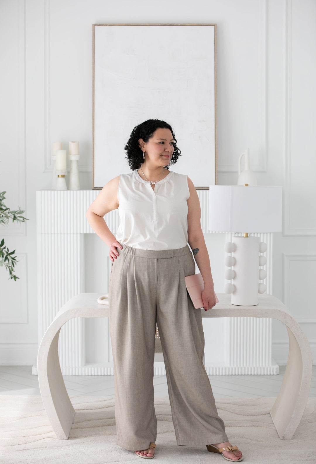 Woman in a white blouse and beige pants standing in a minimalistic room with a white wall and lamp.