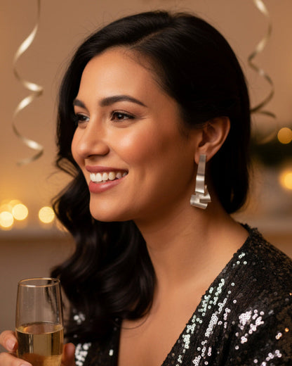 Woman wearing bold silver earrings holding a glass of champagne with party decorations in the background (AI)