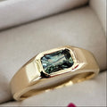 A yellow gold ring with a rectangular green gemstone, presented in an open light gray jewelry box.