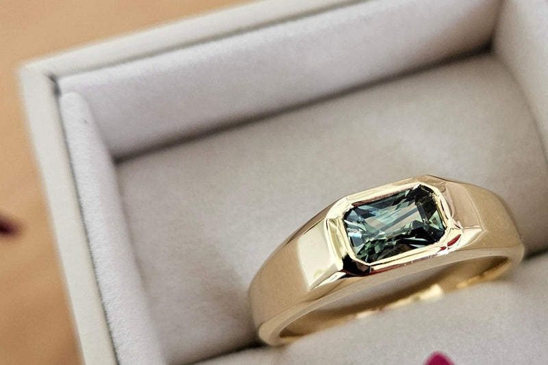 A yellow gold ring with a rectangular green gemstone, presented in an open light gray jewelry box.