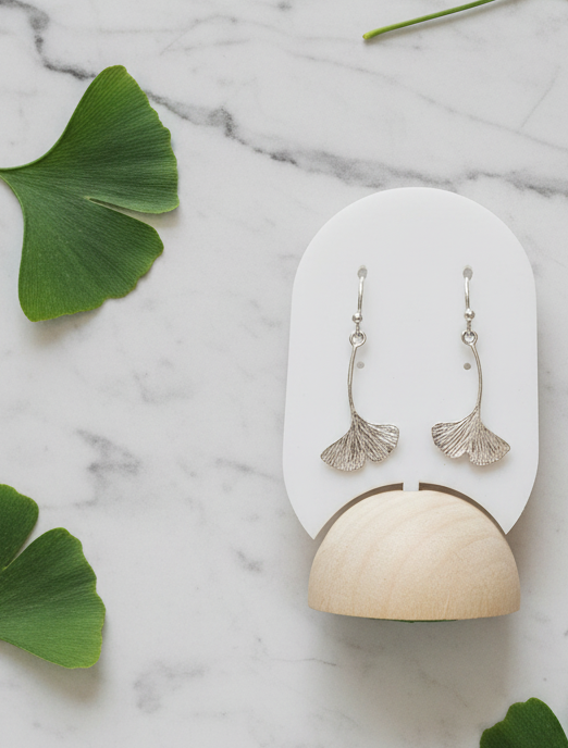 Silver ginkgo leaf earrings on a white and beige stand with green leaves on a marble background