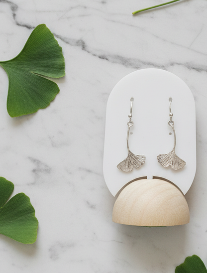 Silver ginkgo leaf earrings on a white and beige stand with green leaves on a marble background