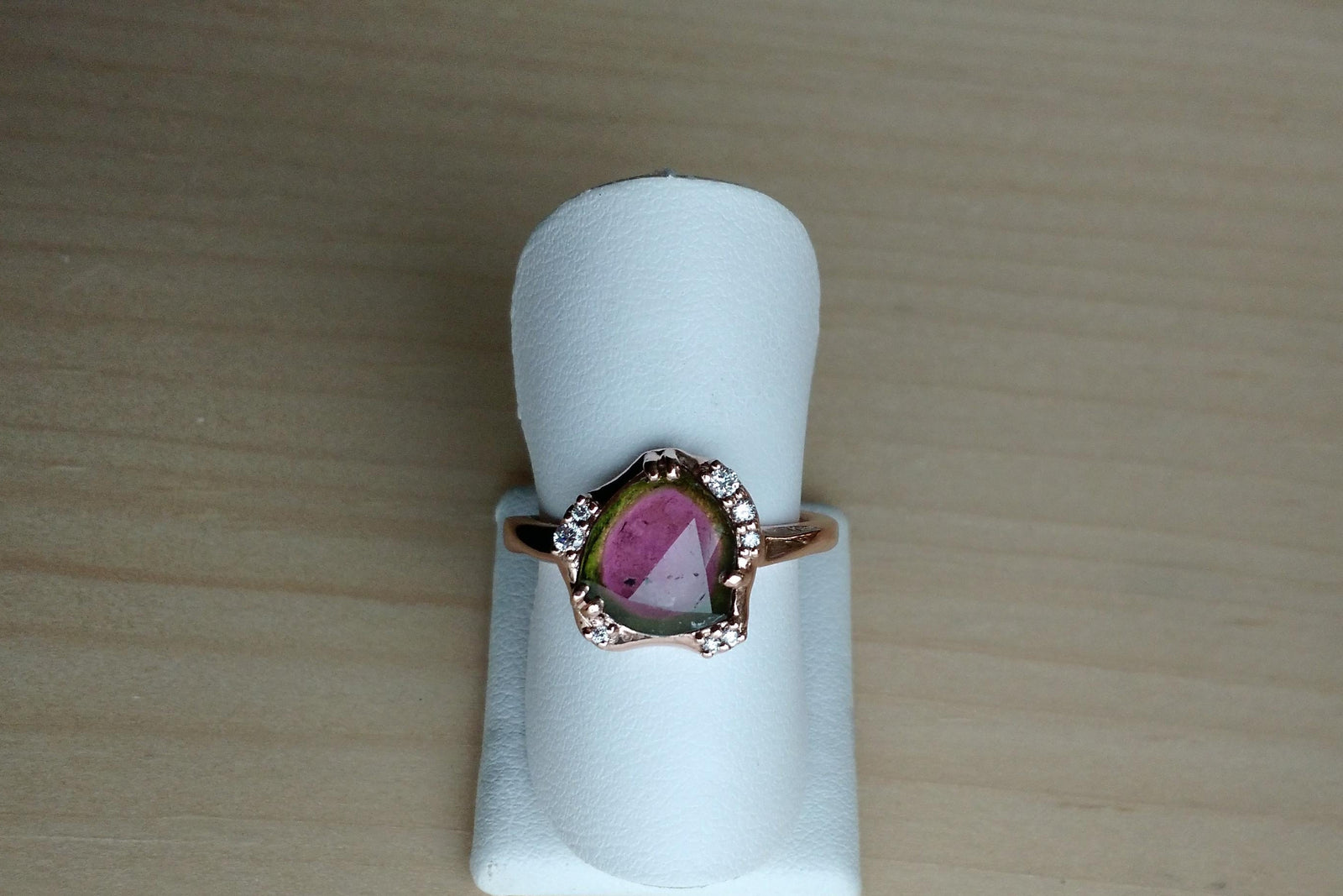 Rose gold ring with a rose cut watermelon tourmaline and diamonds on a white stand against a wooden background