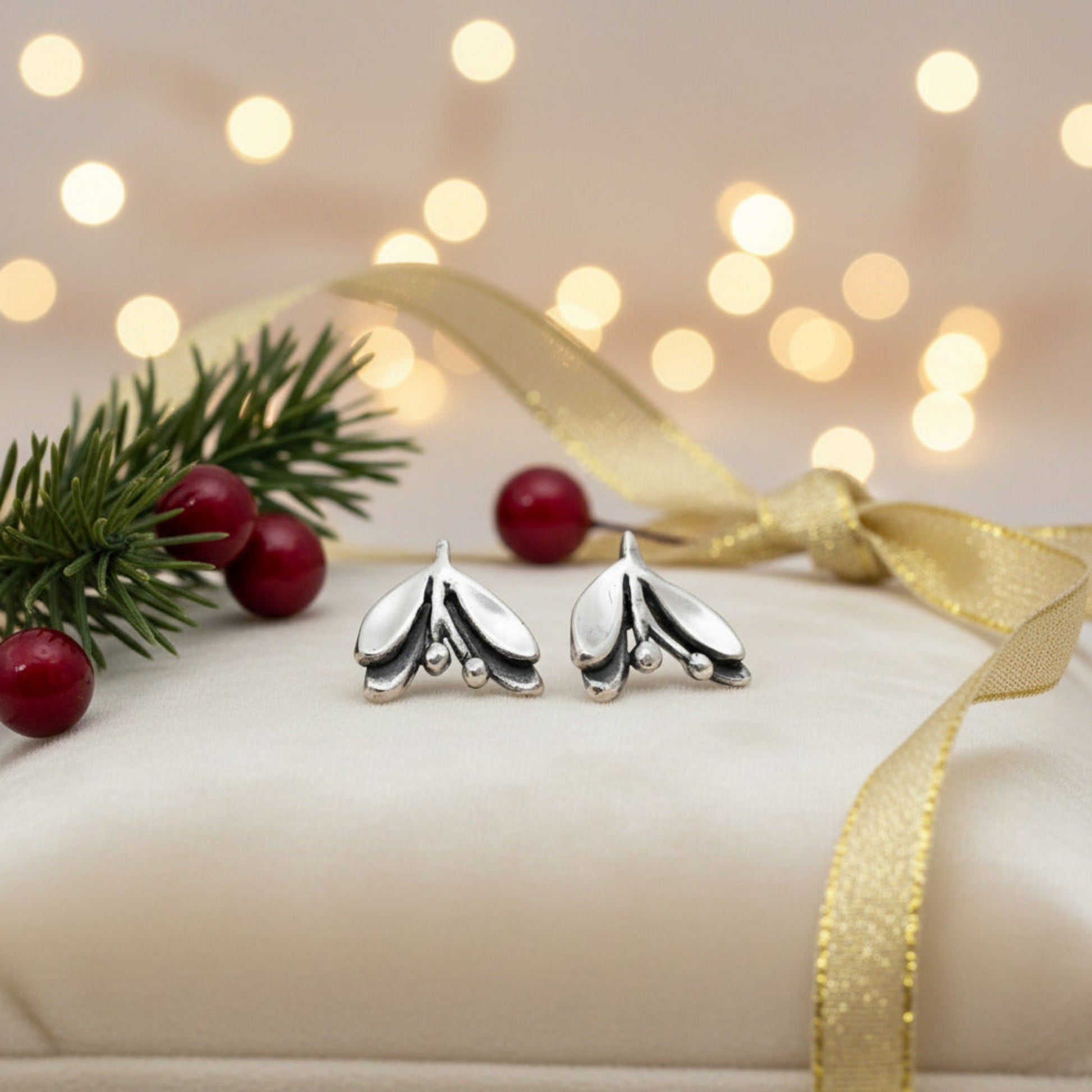 Silver mistletoe earrings on a white cushion with pine branches, berries, blurred lights in the background