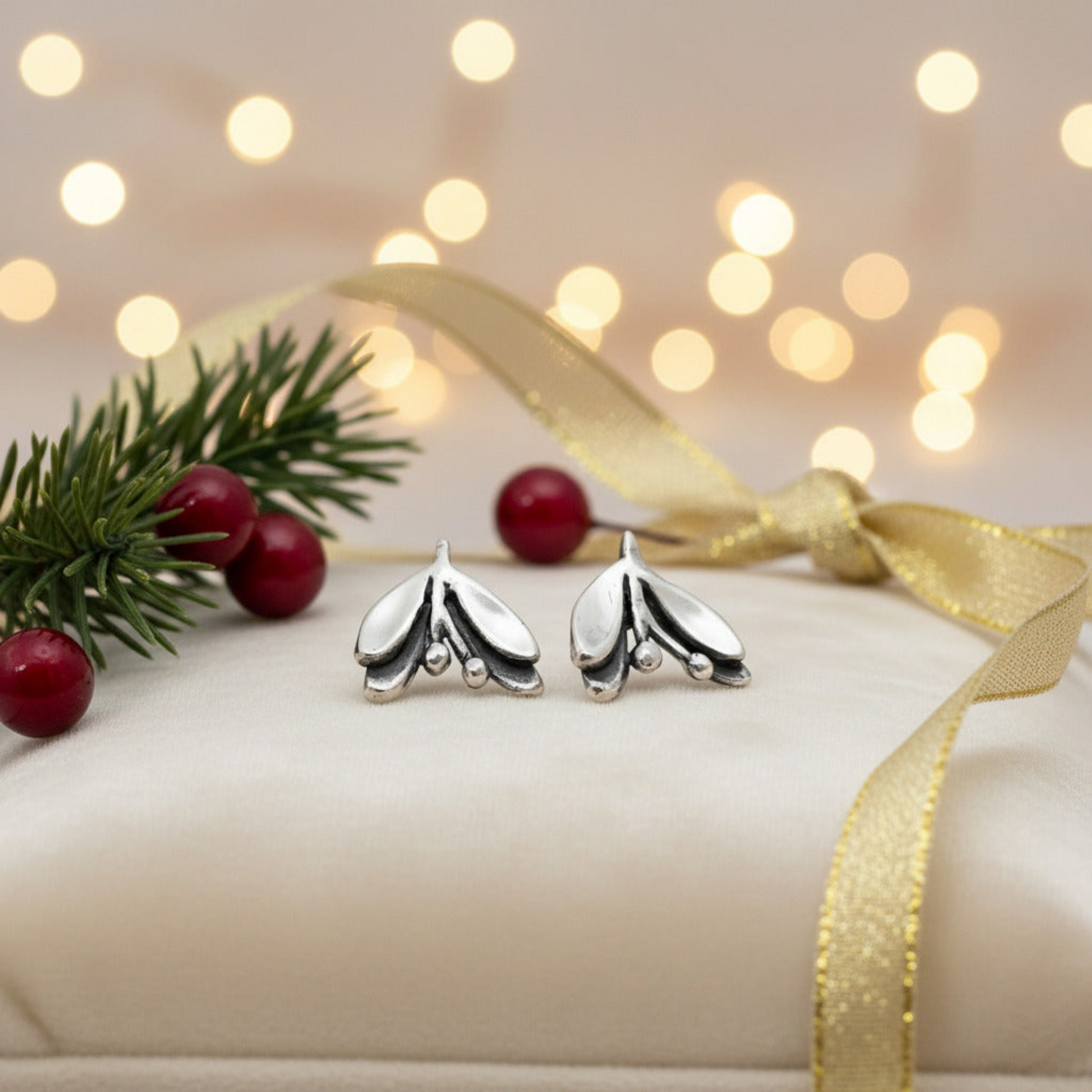 Silver mistletoe earrings on a white cushion with pine branches, berries, blurred lights in the background