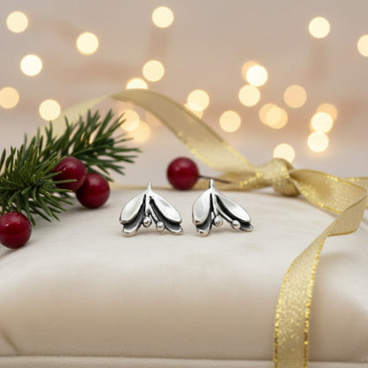 Silver mistletoe earrings on a white cushion with pine branches, berries, blurred lights in the background