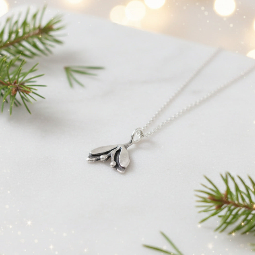 Silver necklace with a mistletoe pendant on a white surface with green branches and blurred lights in the background