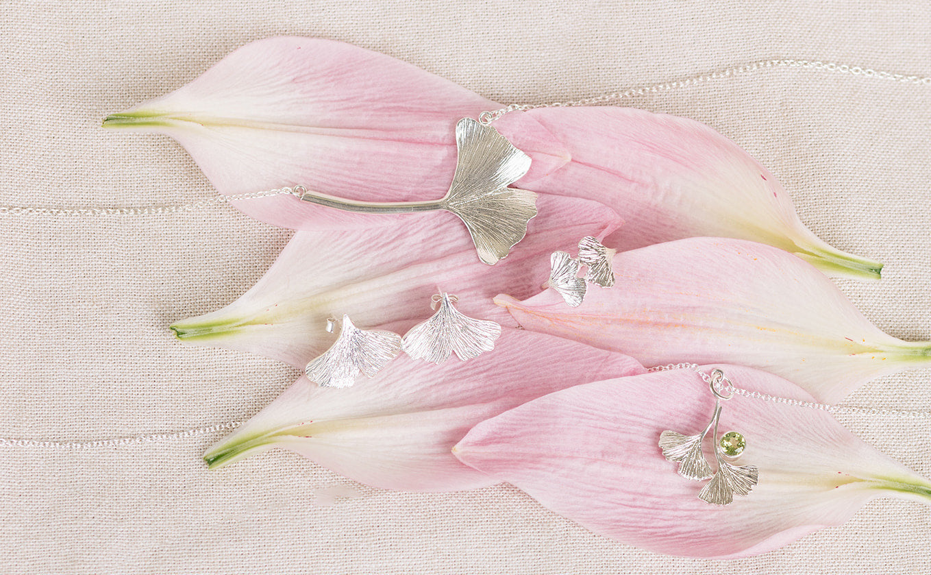 Silver ginkgo jewellery collection resting on pink flower petals with a beige fabric background