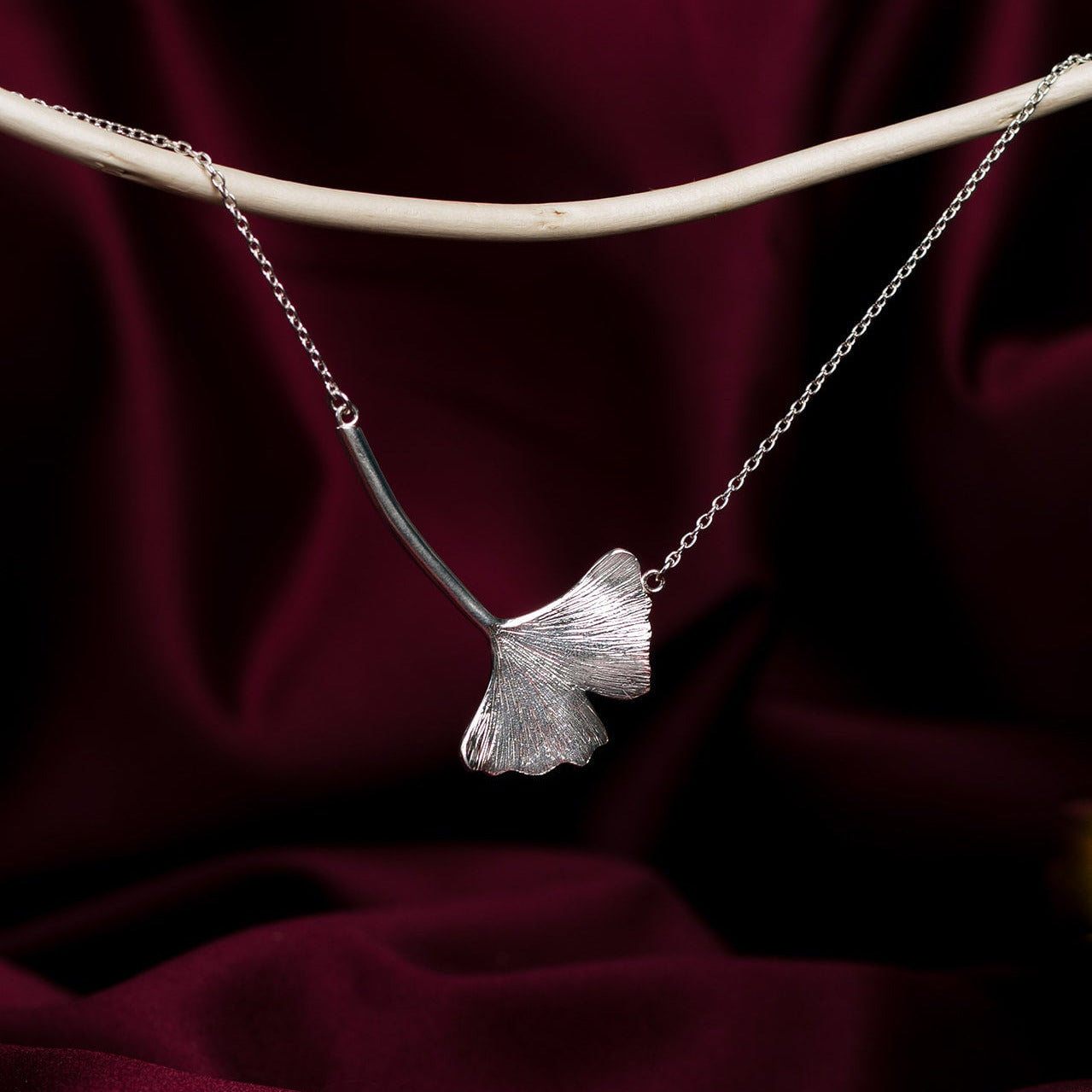 Silver ginkgo leaf-shaped pendant on a chain against a dark red fabric background