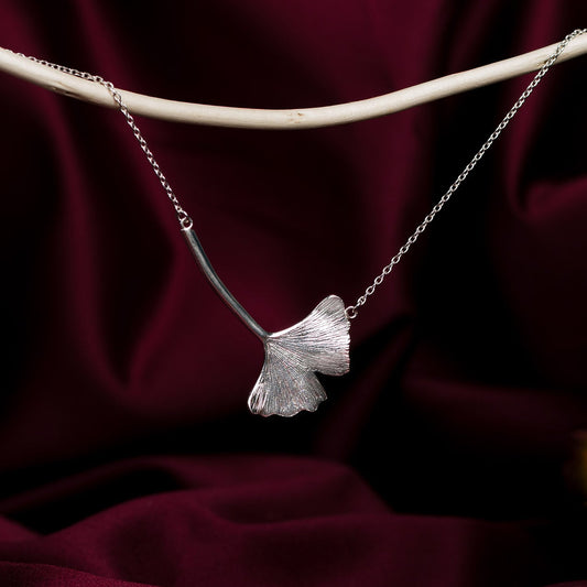 Silver ginkgo leaf-shaped pendant on a chain against a dark red fabric background