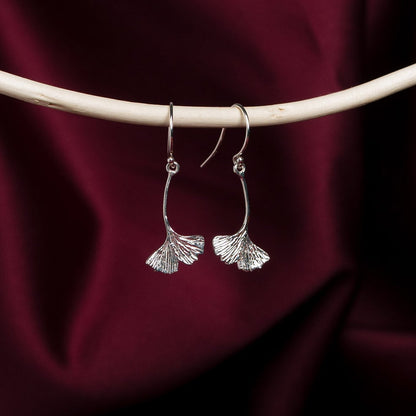 Silver ginkgo leaf earrings on a dark red fabric background