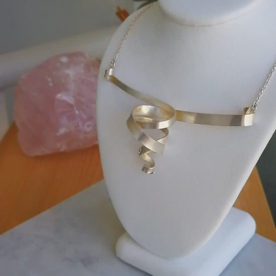 A video showing a sterling silver double spiral pendant necklace with a matte finish and silver chain on jewellery stand beside a potted succulent.