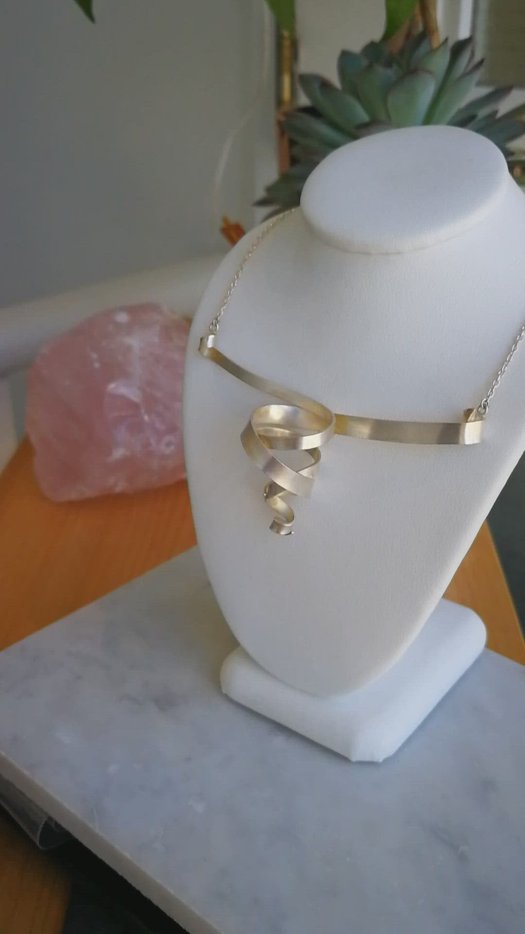 A video showing a sterling silver double spiral pendant necklace with a matte finish and silver chain on jewellery stand beside a potted succulent.