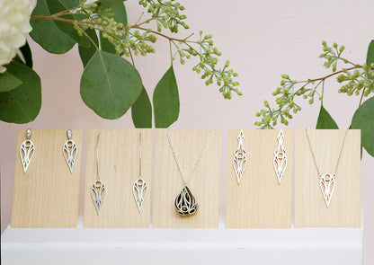 A collection of sterling silver geometric art deco teardrop and dagger jewellery on pale wood panels. From left to right: teardrop stud earrings, teardrop hoop earrings, fig pendant necklace, dagger stud earrings, and teardrop pendant necklace. With flowers and leaves behind.