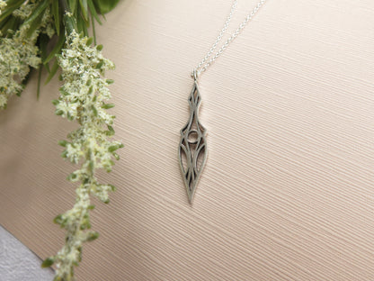Art deco dagger pendant necklace beside white flowers on a beige textured background.