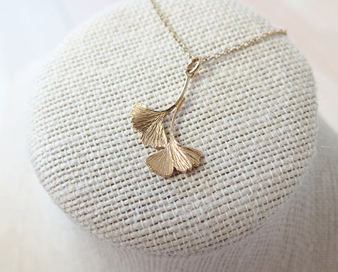 A gold double ginkgo leaf pendant necklace. Assymetrical and textured with a bright shiny finish. The pendant and gold chain are resting on a fabric pedestal. 