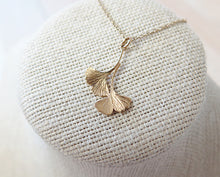 Load image into Gallery viewer, A gold double ginkgo leaf pendant necklace. Assymetrical and textured with a bright shiny finish. The pendant and gold chain are resting on a fabric pedestal. 
