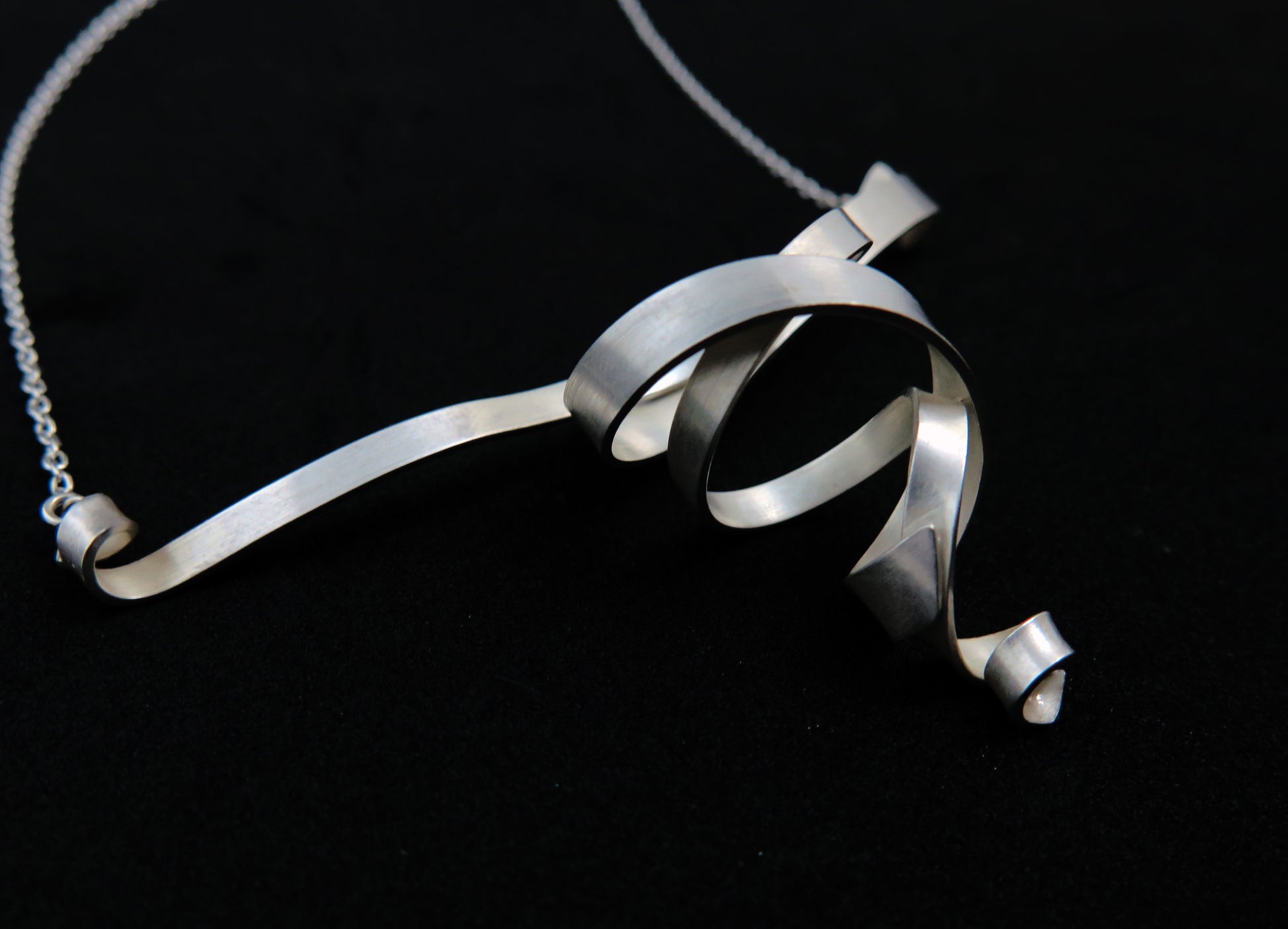 A sterling silver sculptural double spiral pendant necklace with a matte finish and silver chain on a black background.