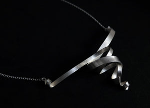 An elegant modern sculptural double whirl necklace with a matte finish on a black background.