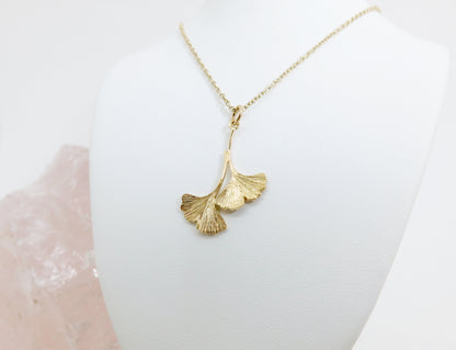 A gold pendant necklace of two assymetrical and organically textured ginkgo leaves with a gold chain on a white jewellery stand.