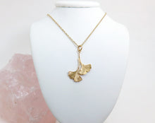 Load image into Gallery viewer, A gold double ginkgo leaf pendant necklace. Assymetrical and textured with a bright shiny finish. On a gold chain hanging on a white jewellery stand.
