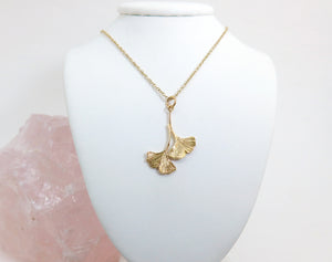 A gold double ginkgo leaf pendant necklace. Assymetrical and textured with a bright shiny finish. On a gold chain hanging on a white jewellery stand.