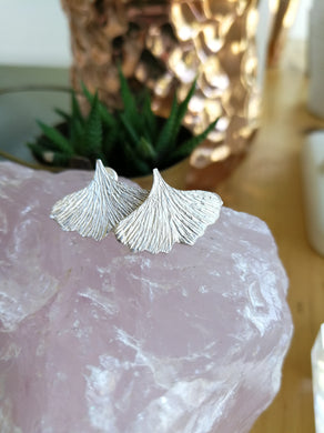 Textured sterling silver ginkgo leaf shaped stud earrings resting on a piece of rose quartz with a plant in the background