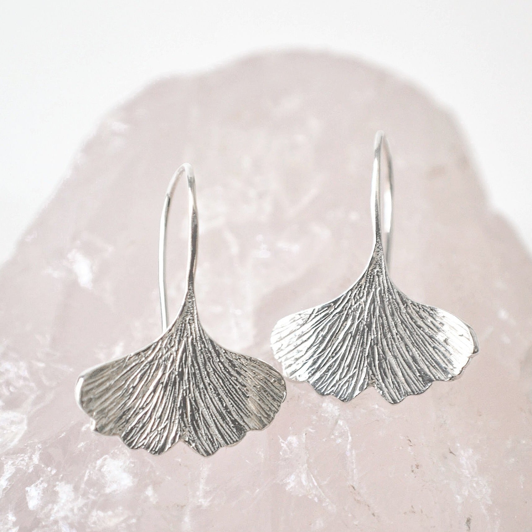 Textured sterling silver ginkgo leaf shaped dangle earrings resting on a piece of rose quartz