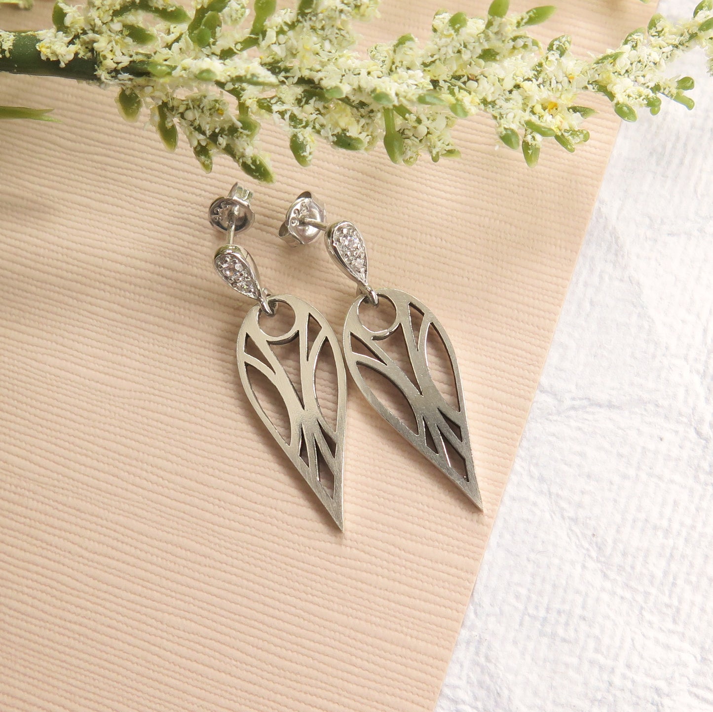 Geometric art deco teardrop stud earrings in sterling silver with cubic zirconia gemstones on a beige and white textured paper with a sprig of flowers.