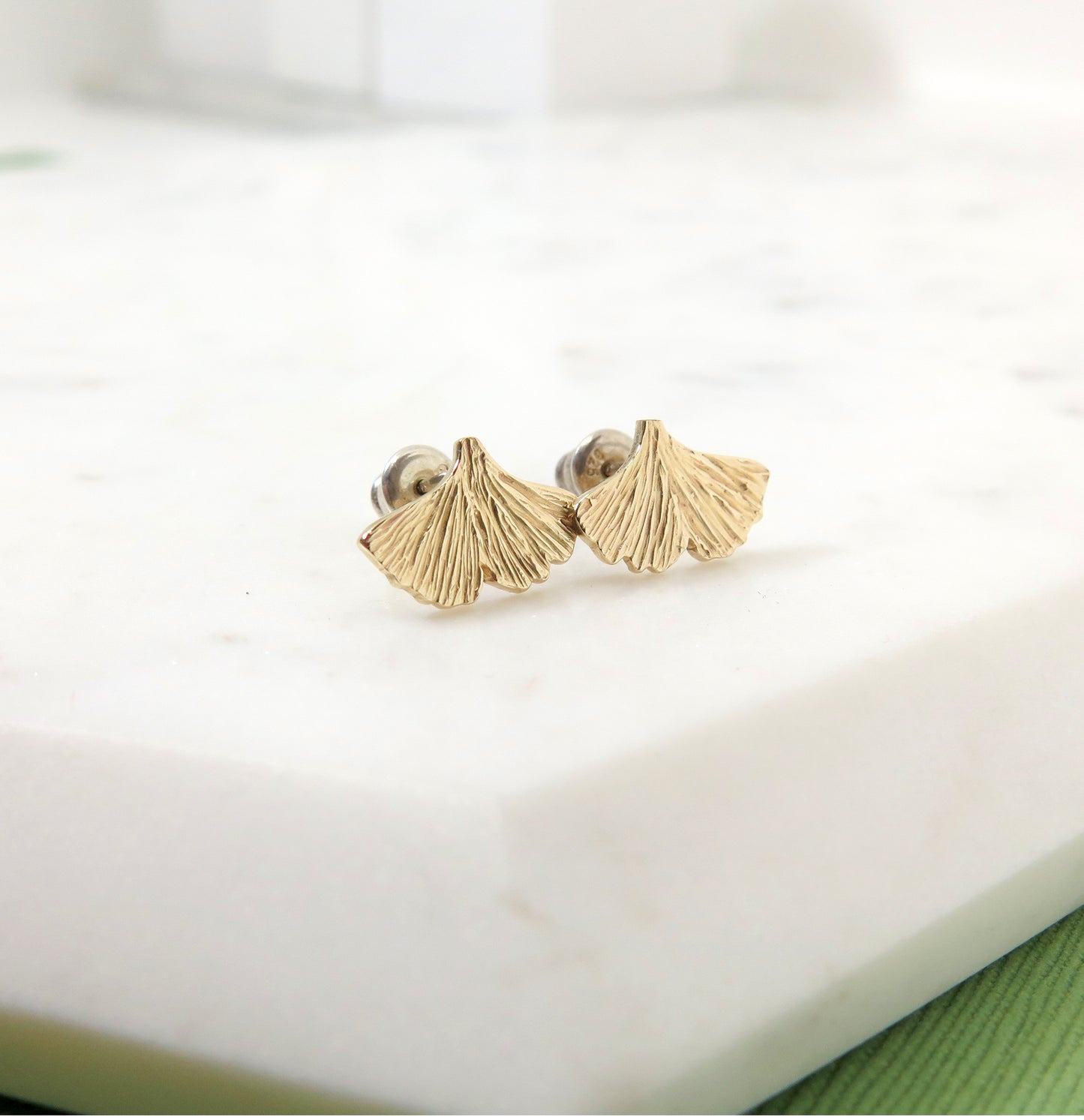 Textured gold ginkgo leaf stud earrings resting on a piece of marble
