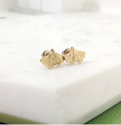 Textured gold ginkgo leaf stud earrings resting on a piece of marble