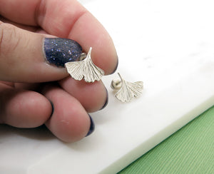 Textured sterling silver ginkgo leaf stud earrings in a hand with nail polish on the nails.